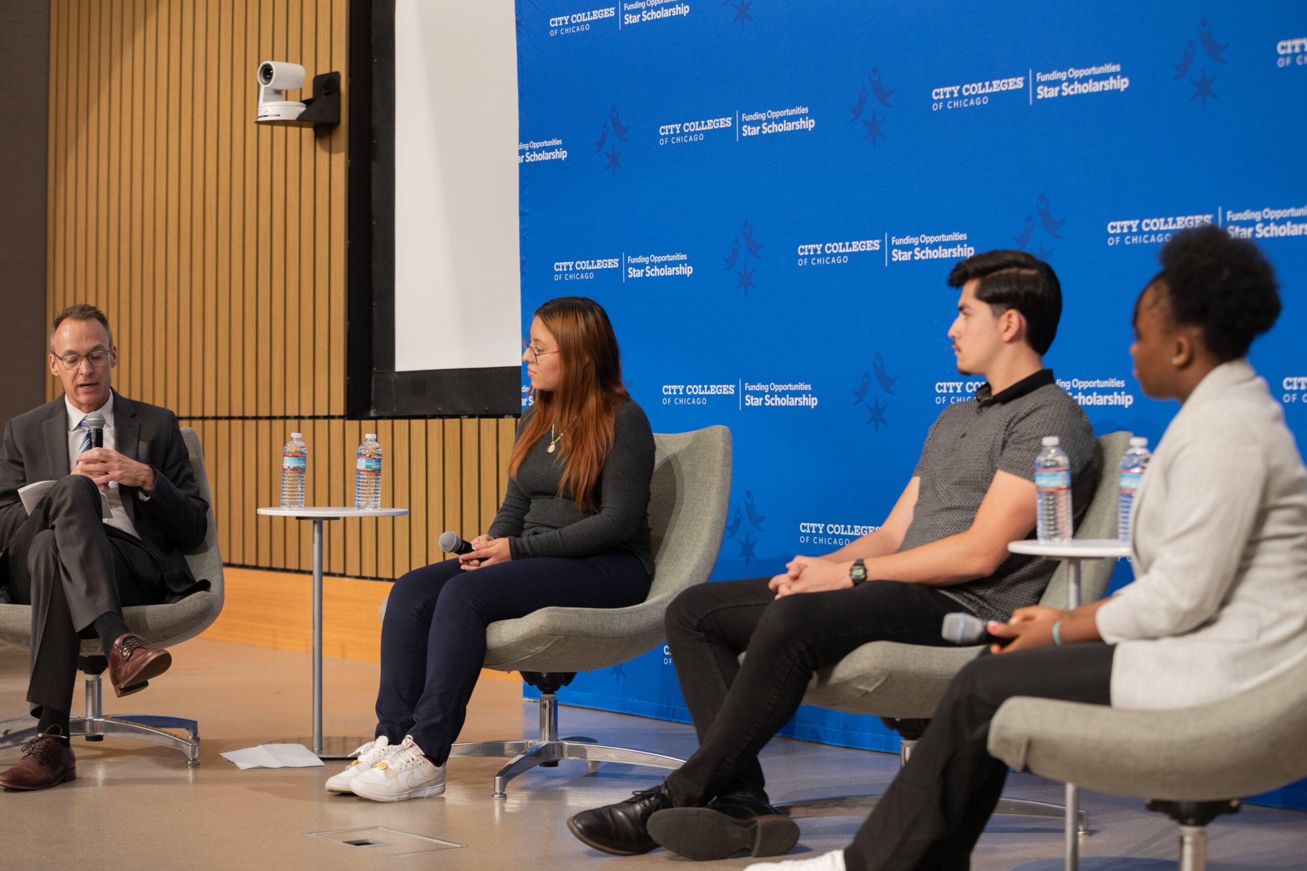Four people sit on stage in front of a blue background and speak on a panel.