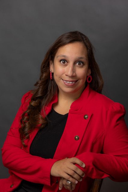 A woman with long dark, lightly curled hair that is tossed over her right shoulder smiles at the camera. She is sitting in a chair, leaning slightly to her left with her right hand over her left. She's wearing a red blazer and a black top.
