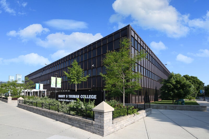 The exterior of a black building with lots of large windows and a sidewalk. The sign outside the building reads Harry S Truman College.